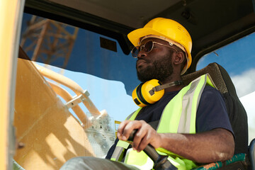Poster - Young black male driver of construction machine in safety helmet and uniform sitting by steer and looking through front window