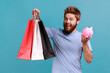 Portrait of smiling bearded man holding shopping bags and piggy bank, cashback from buying purchases, expressing positive emotions. Indoor studio shot isolated on blue background.