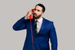 Pensive bearded man holding phone handset, having conversation, looking away with thoughtful expression, wearing official style suit. Indoor studio shot isolated on gray background.