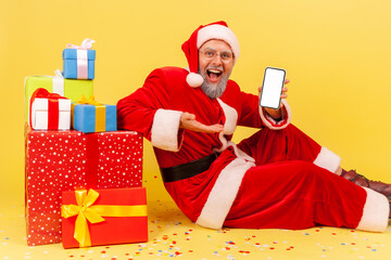 Wall Mural - happy elderly man with gray beard wearing santa claus costume presenting blank white display of cell
