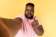 Portrait of serious aggressive man wearing pink shirt taking selfie, showing rude gesture with middle finger, point of view photo, POV. Indoor studio shot isolated on yellow background.