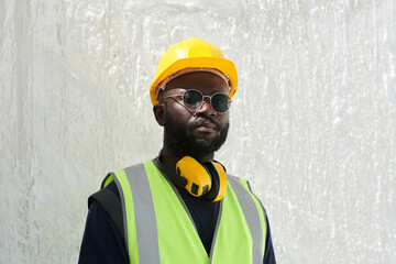 Poster - Young serious African American male engineer in safety helmet, reflective vest and eyeglasses looking at camera against white marble wall