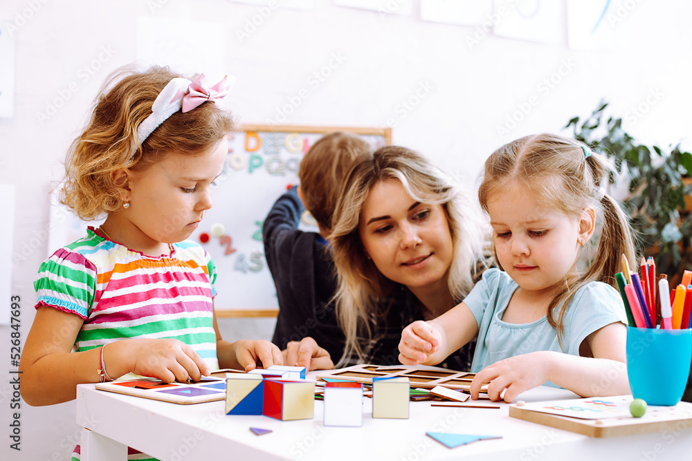 Beautiful little girls with interest fold colored cubes on desk in playroom of kindergarten. Young educator sitting with children at table on floor and watch play. Lesson in daycare centre