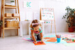© Татьяна Волкова - Pretty little girl sitting bent over on knees on floor in playroom and draw picture on red sheet of paper with marker. Kindergartener develop artistic skills in daycare. Hobby, childish fantasy