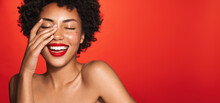 Beauty And Spa. Happy Laughing African American Woman, Wearing Red Lipstick, Touches Her Glowing Nourished Skin With Cosmetic Product On, Standing Over Red Background
