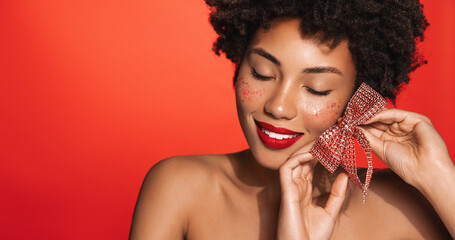 Wall Mural - Portrait of smiling Black female model with glitter, looking down sensual, showing bowtie, posing against red background