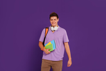 Young happy man student in casual clothes posing with backpack and notebooks, isolated on studio violet background. Education concept.