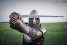 The Knight In The Plate Armor And Helmet Holds A Sword In Hands Close Up.
