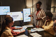 © Seventyfour - Portrait of young black woman giving presentation on marketing to team in dimly lit office