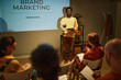 © Seventyfour - Portrait of young black man giving presentation on brand marketing to team while standing by projector screen and addressing audience in business training