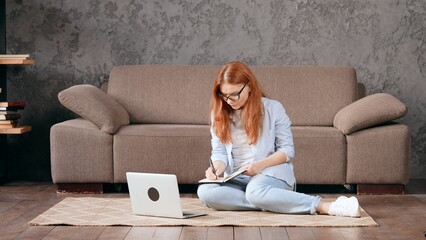 Sticker - Caucasian red-haired woman sitting on the carpet while taking notes in a notebook.