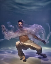 Young Man In White Ribbons And Trousers Underwater In The Pool On A Dark Background
