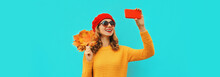 Autumn Portrait Of Happy Smiling Young Woman Taking Selfie With Smartphone With Yellow Maple Leaves Wearing Sweater, French Beret On Blue Background