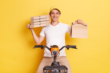 Indoor shot of delivery man riding bike and holding paper packages with takeaway food, wearing t-shirt and cap, looking at camera with smile, isolated over yellow background.