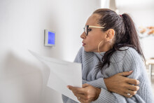 Woman With Papers In Her Hands Shivers In Sweater Looking At The Thermostat On The Wall