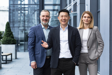 Successful diverse business team, three workers smiling and looking at camera, dream team with asian boss outside office building, colleagues in business suits