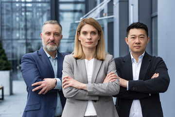 Successful and serious diverse team of three business people, man and woman focused looking at camera with arms crossed, portrait of co-workers outside office building