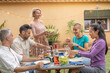 © deliris - Happy friends laughing with big smile around the table in a summer diner. Woman serves the food at house patio. Smiles, cheerfulness concept.