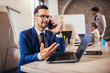 © Mediteraneo - Happy businessman using laptop and phone with team discussing project in the background.