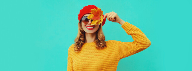 Autumn portrait of happy smiling young woman with yellow maple leaves wearing knitted sweater, red beret on blue background