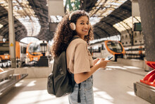 Nice Young African Girl Looking At Camera Uses Her Smartphone Stands On Subway Station In Sunny Day. Brunette With Curly Hair Wears Casual Style Of Clothes. Concept Tourist, Vacation.