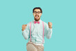 Cheerful happy male hipster nerd rejoices in his success standing on light blue background. Guy with mustache and beard wearing shirt, suspenders and glasses happily clenched his fists shouting YES.