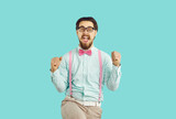Fototapeta  - Cheerful happy male hipster nerd rejoices in his success standing on light blue background. Guy with mustache and beard wearing shirt, suspenders and glasses happily clenched his fists shouting YES.