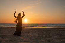 Silhouette Of A Woman In Long Black Dress Dancing On The Beach Near Ocean At Sunset
