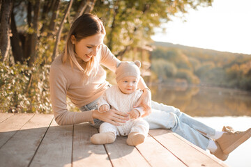 Smiling young woman holding playing with baby boy 1 year old wear knit clothes over nature background and lake with forest. Autumn season. Motherhood.