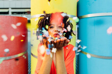 Smiling Young Woman Blowing Confetti In Front Of Colorful Pipes
