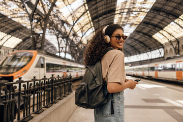 Wall Mural - Positive young african woman with headphones walking along subway station with her smartphone. Brunette wears casual clothes, sunglasses and backpack. Concept of use.