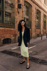 Wall Mural - In full length young african woman in sunglasses looking at camera stands on street alone. Brunette wears dress and jacket. Concept stylish vacation.