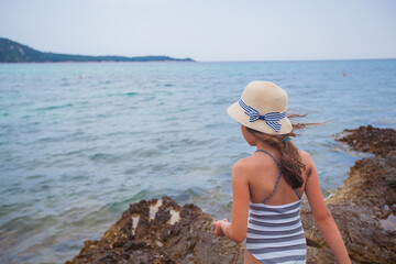 Wall Mural - Summer smiling portrait of little girl with straw hat at rocks shore. Adorable child on vacation. Seascape in background. 