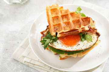 Wall Mural - Savory breakfast - fresh homemade belgian waffle sandwich with green lettuce, crisp bacon and fried egg on white plate on beige colored table cloth, glass with lemon water, cup of coffee