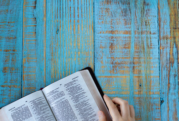 Wall Mural - Human hand holding an open Holy Bible Book on blue wooden background with copy space. Top table view. Study and read Christian Scripture, biblical concept.