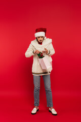 Canvas Print - full length of young woman in sweater and santa hat putting gift box with bow into paper bag on red.