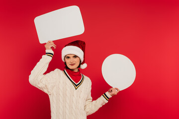 Wall Mural - smiling young woman in santa hat and sweater holding speech bubbles isolated on red.