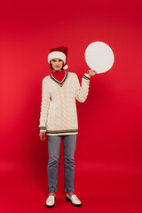 Wall Mural - full length of young woman in santa hat and sweater holding speech bubble on red.