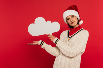 Wall Mural - young woman in santa hat and sweater holding thought bubble and pointing with hand isolated on red.