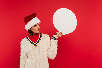 Wall Mural - young woman in santa hat and sweater holding speech bubble isolated on red.