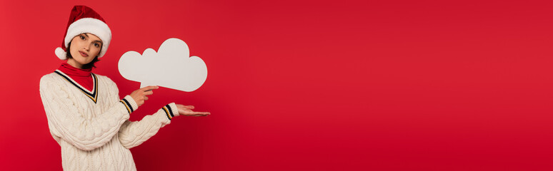 Wall Mural - young woman in santa hat and sweater holding thought bubble and pointing with hand isolated on red, 