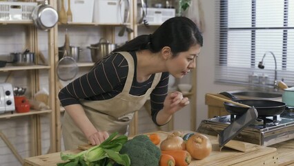 Wall Mural - asian new wife leaning on the kitchen table and using a touchpad to learn cooking by watching online culinary tutorials in the kitchen at home during daytime.