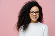 Headshot of charming young female with Afro hairstyle, smiles pleasantly at camera, feels very happy, wears transparent glasses and white jumper, isolated on rosy background. Amused ethnic female