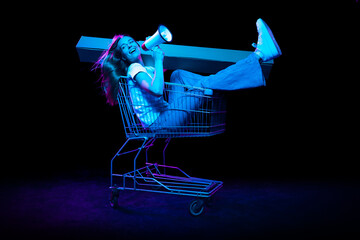 Cheerful astonished girl sitting in shopping cart and inviting for shopping, black friday, seasonal sales isolated over dark background in neon light.