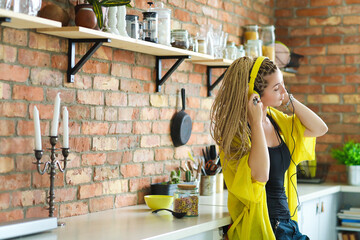 Wall Mural - Photo of bright,cool,beautiful woman with dreadlocks enjoying music in yellow headphones while standing in the kitchen.