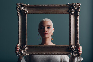 Wall Mural - Millenial young woman with short blonde hair holds gilded picture frame in hands behind her face portrait