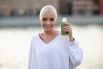 Wall Mural - Portrait of young smiling millenial european short haired woman with green smoothie. Beautiful happy blonde girl outdoor.