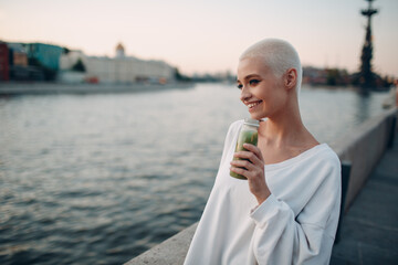 Wall Mural - Portrait of young smiling millenial european short haired woman with green smoothie. Beautiful happy blonde girl outdoor.