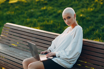 Wall Mural - Portrait of young smiling millenial european short haired woman using laptop at bench on green grass meadow in park. Beautiful happy blonde girl outdoor.