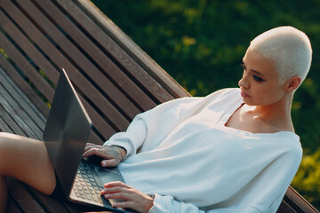 Wall Mural - Portrait of young smiling millenial european short haired woman using laptop at bench on green grass meadow in park. Beautiful happy blonde girl outdoor.